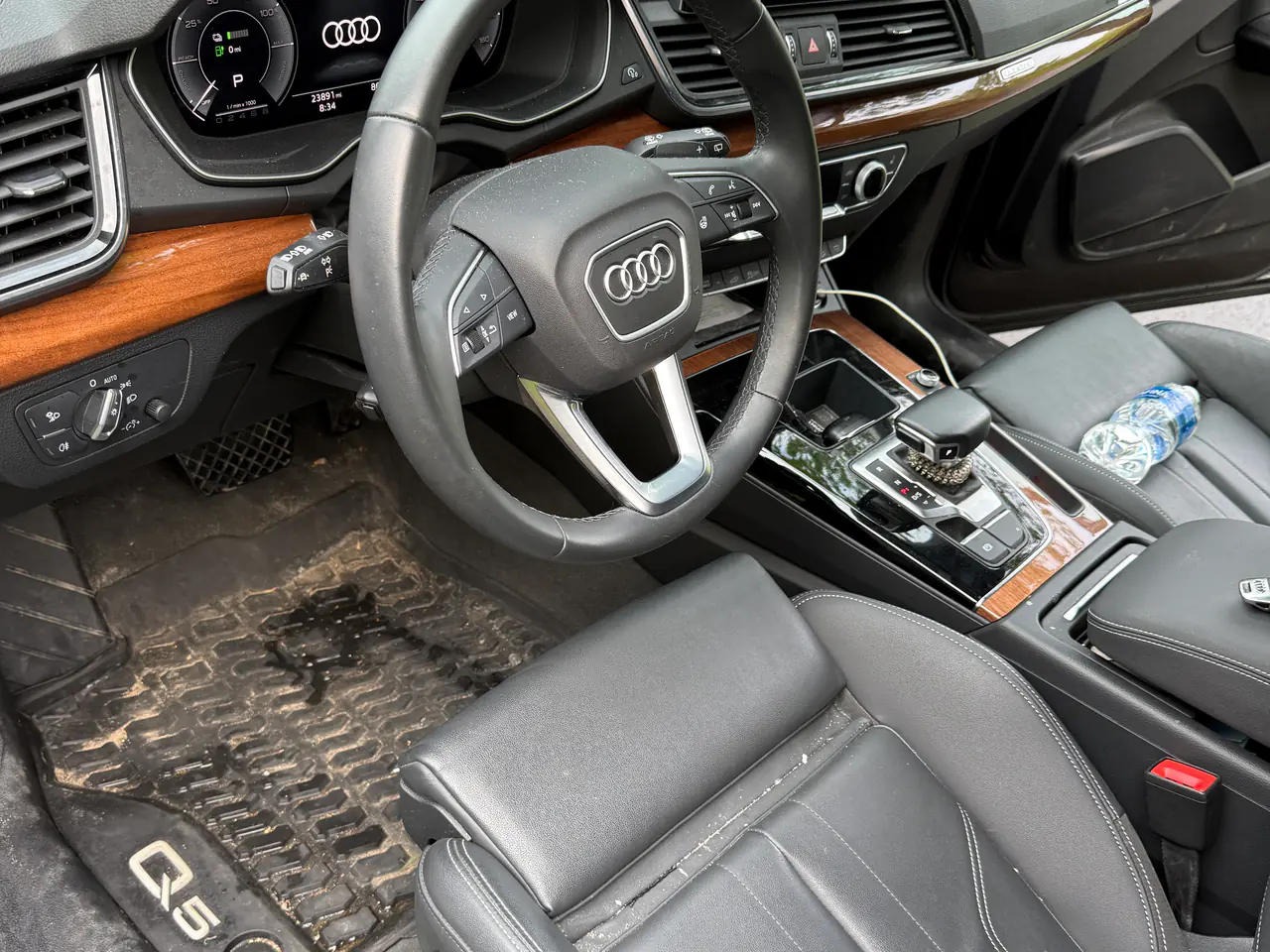 Before an interior detail: an Audi Q5's driver's side, its floor mat caked in mud and a water bottle and dust on the console