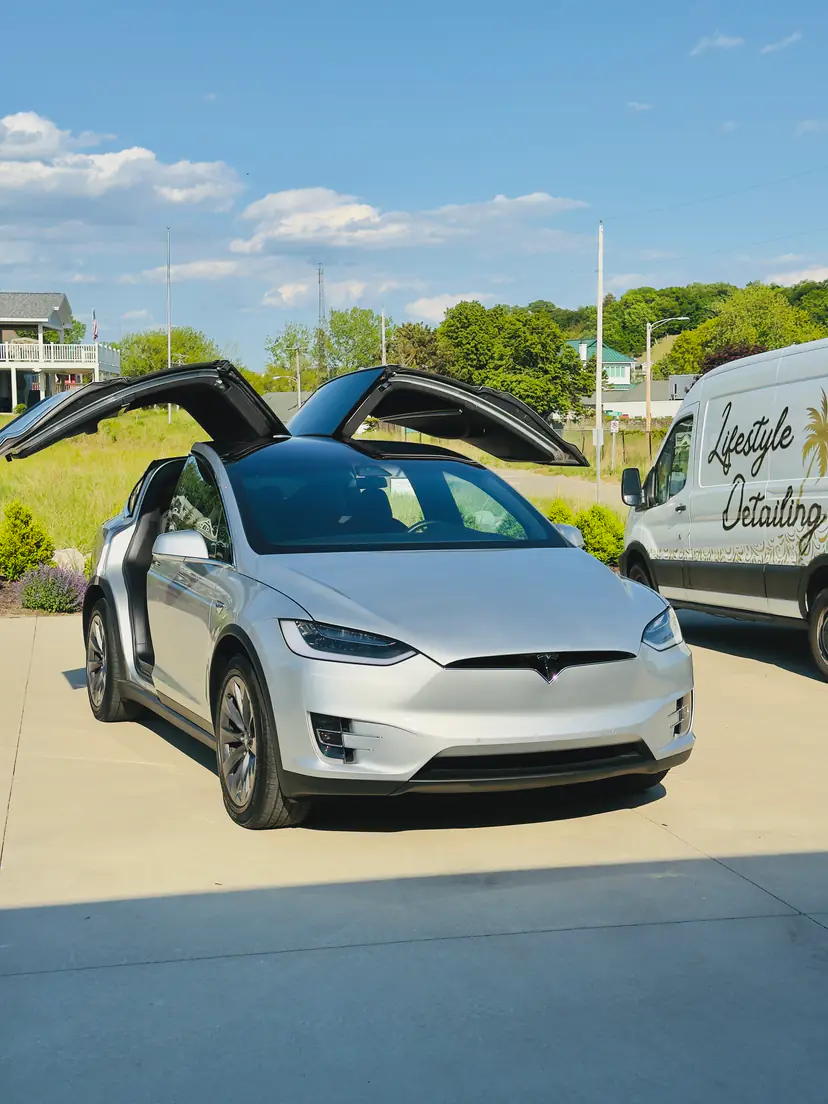 Silver Tesla Model X with its falcon-wing doors raised during a mobile detail, parked beside the Lifestyle Detailing van