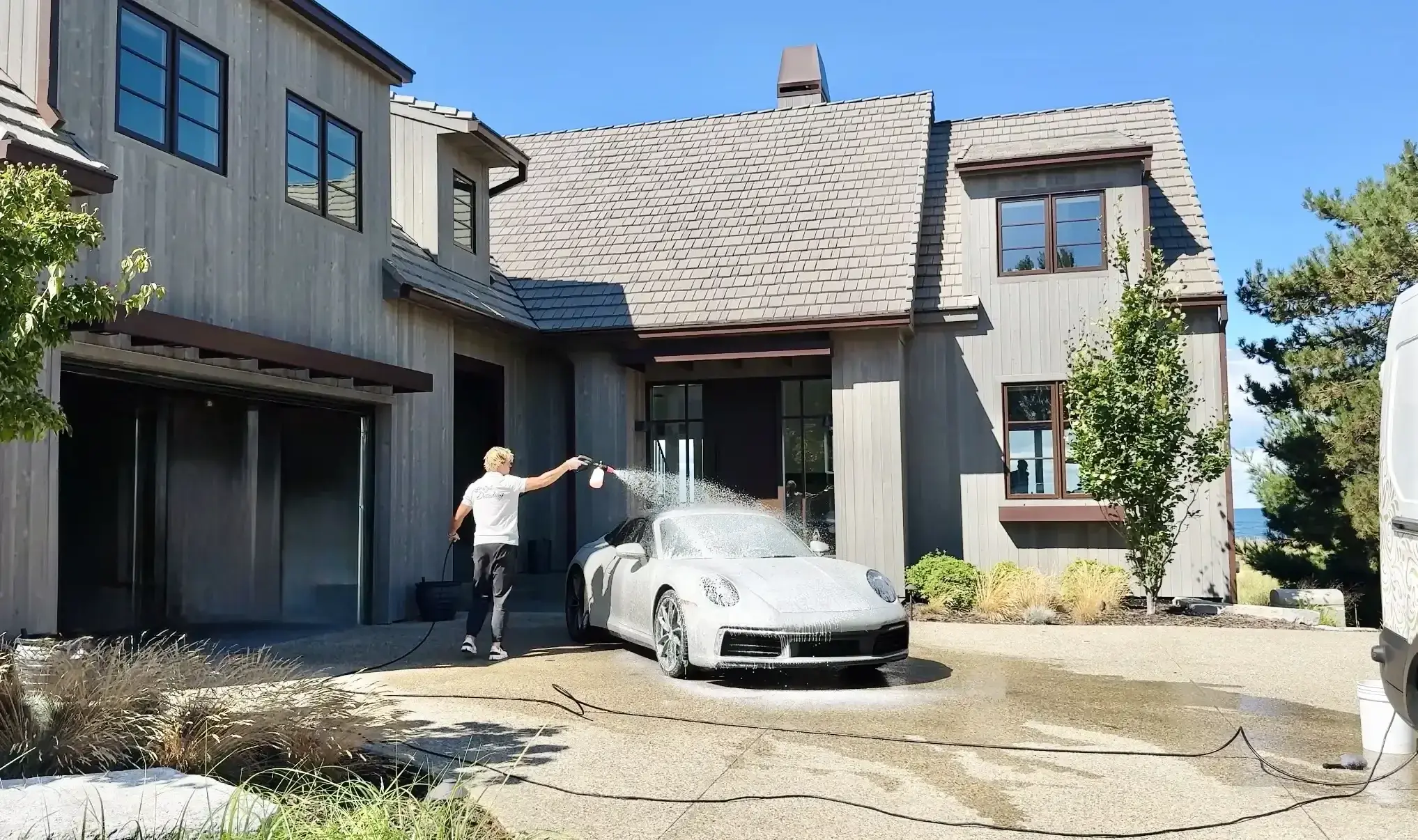 Chase Shooltz rinsing a white Porsche 911 during a mobile detail in the driveway of a modern grey home
