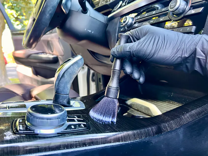 Gloved hand working a soft detailing brush around a BMW's iDrive controller during a mobile interior detail in Grand Rapids Township, Michigan