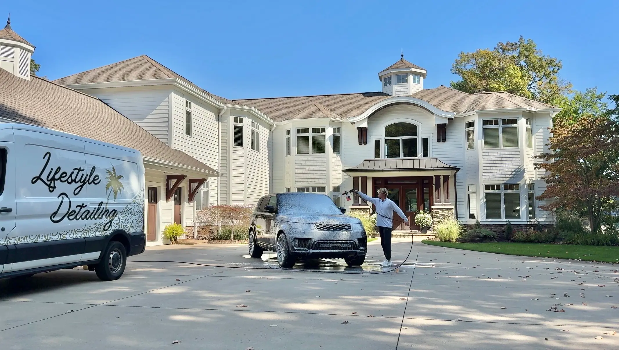 Chase Shooltz rinsing a Range Rover Sport during a mobile hand wash at a large white home, the Lifestyle Detailing van beside it
